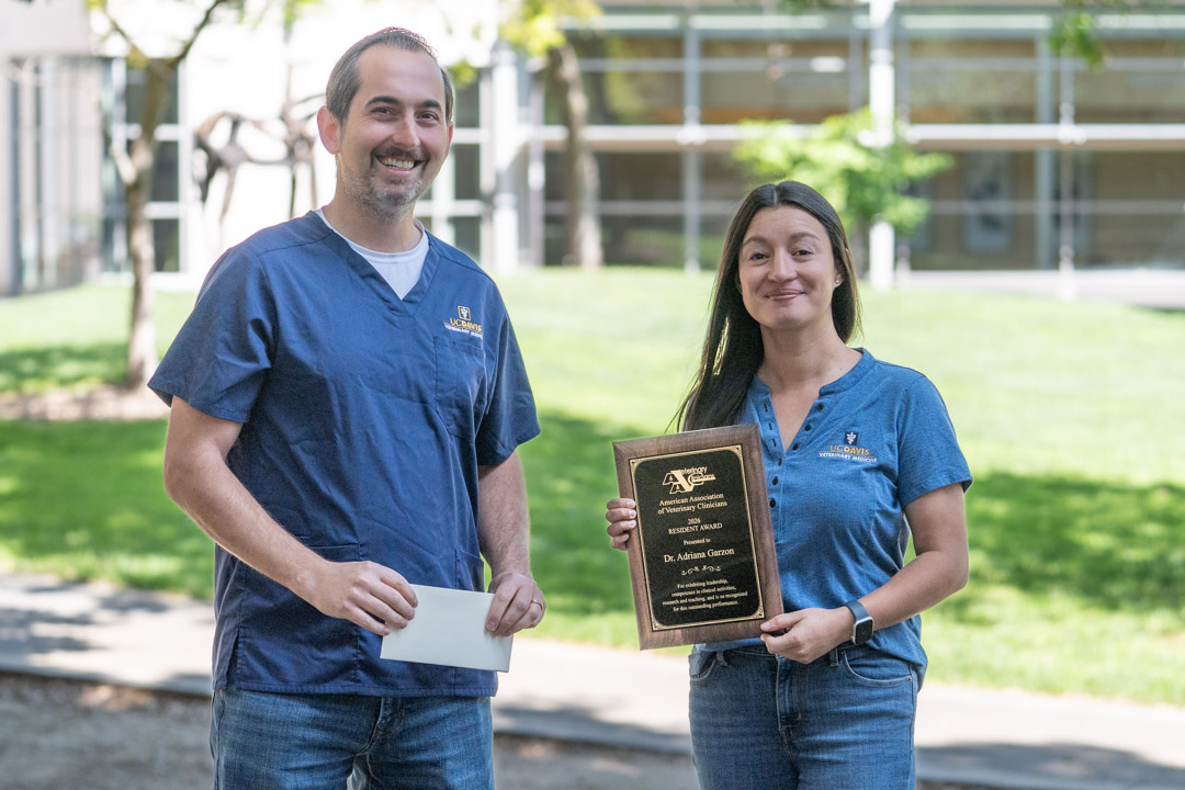 Two smiling colleagues in blue shirts outdoors; woman holds award plaque, man holds envelope