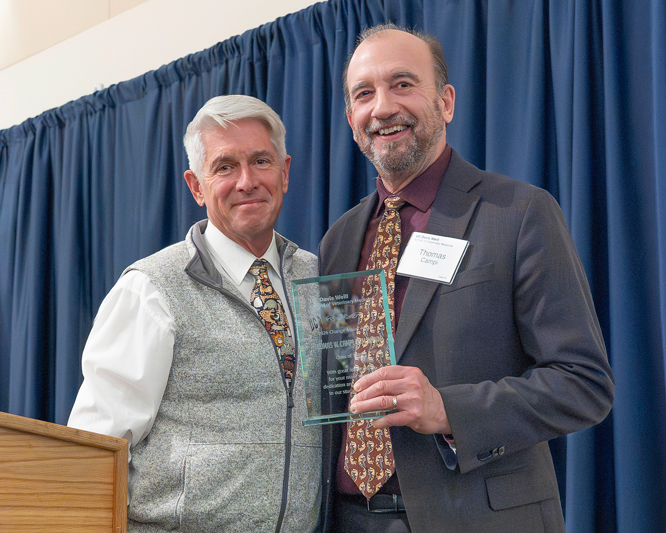 Two men are smiling and posing together, holding an award against a blue curtain backdrop.