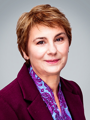 A woman with short brown hair smiles warmly, wearing a purple blazer and colorful scarf.
