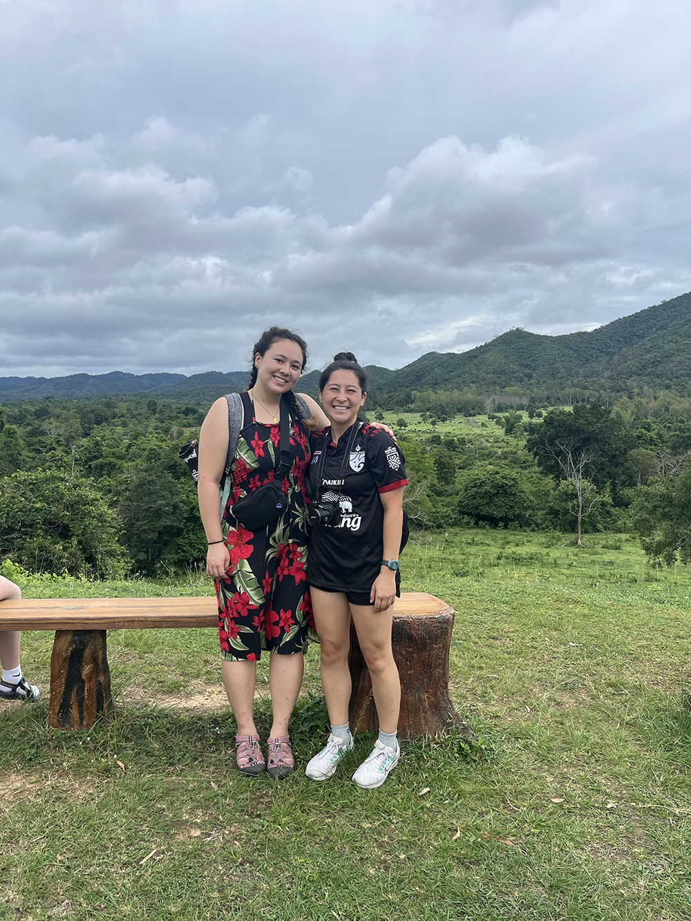 Two women stand together smiling in a lush green landscape with mountains in the background.