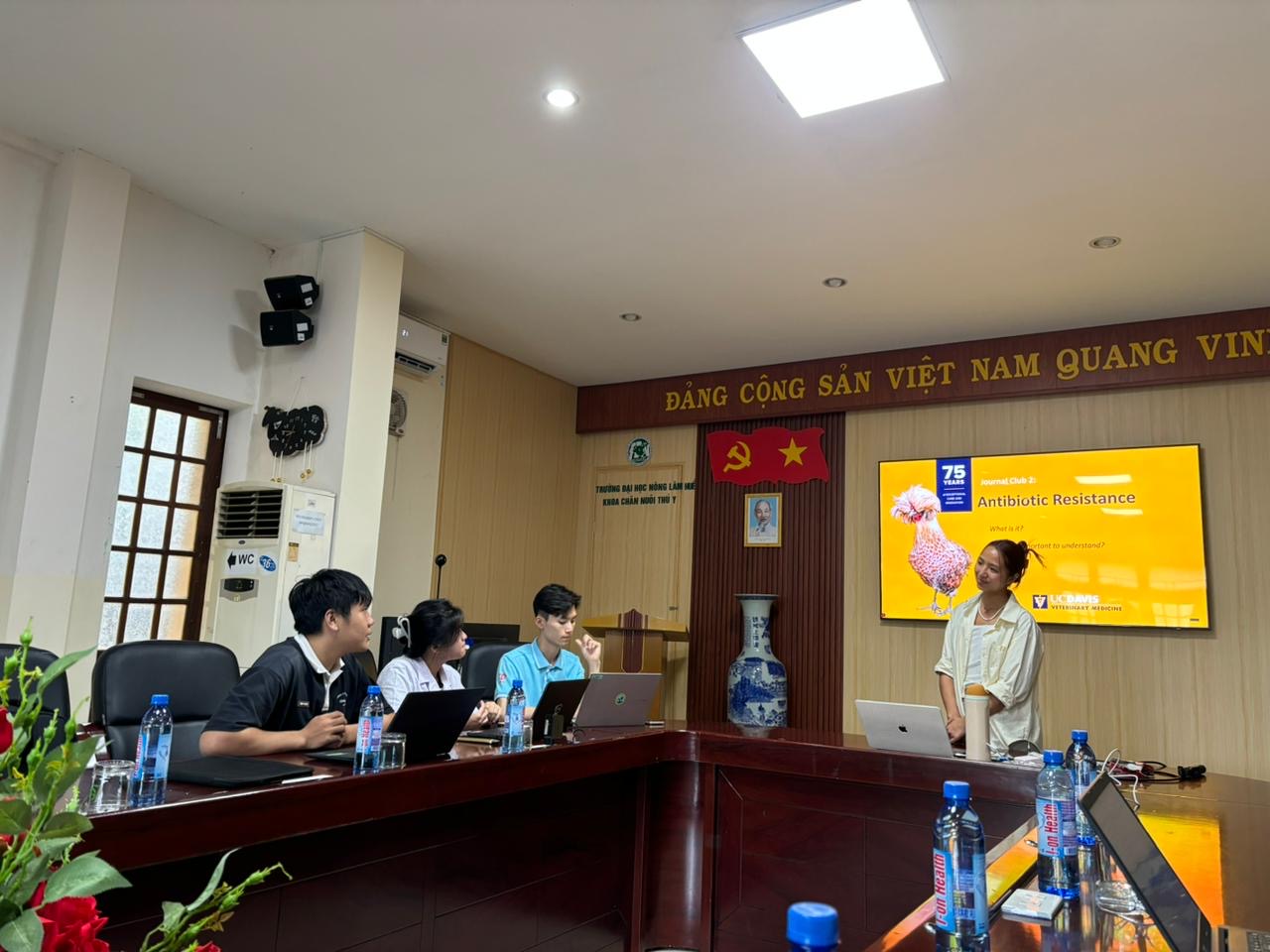 Nhung giving a presentation to 3 people with a slide title, "Antibiotic Resistance" projected on a screen.