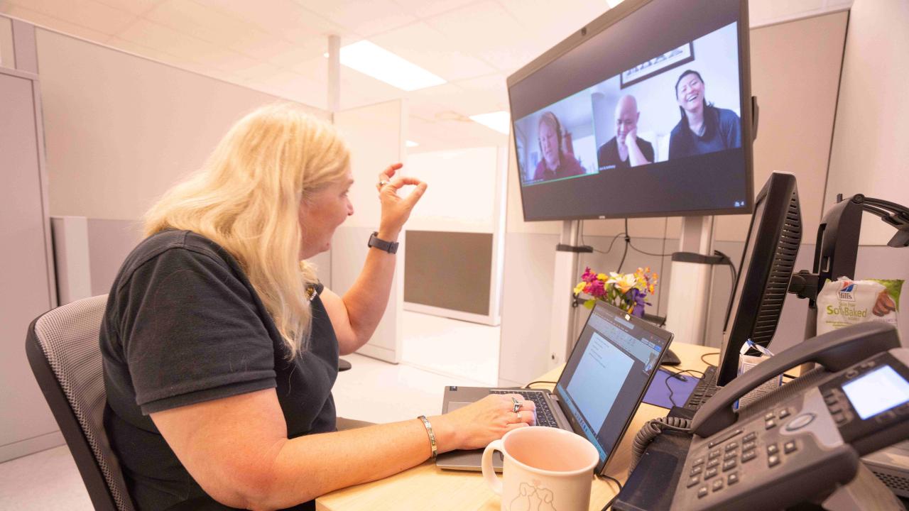 Woman in office cubicle gesturing during video conference on desktop monitor