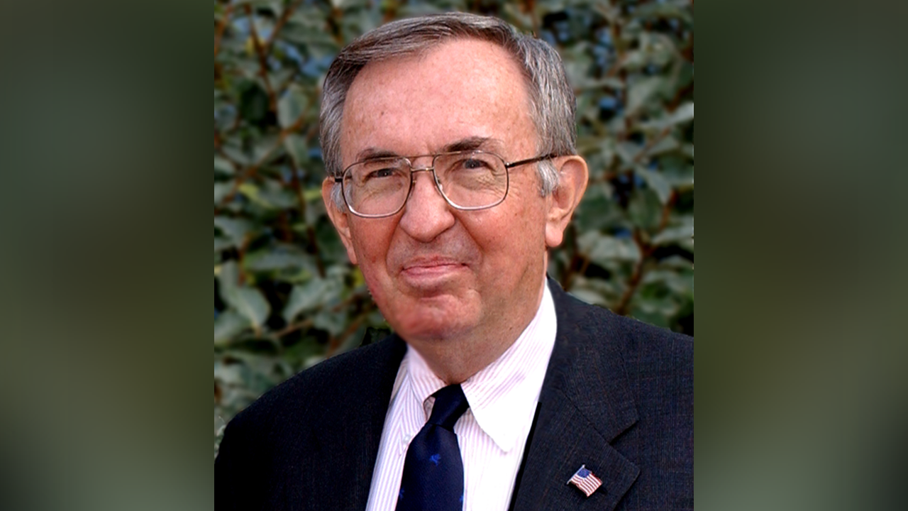 Portrait of an older man in suit and glasses, American flag lapel pin, leafy background.