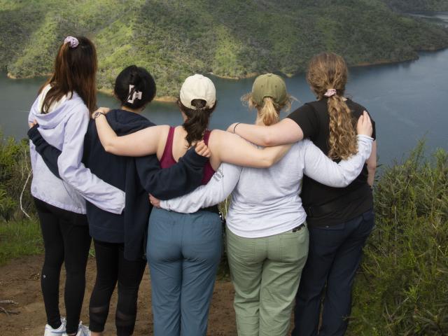 A group of people standing with arms wrapped around each other at the top of a summit overlooking a lake.
