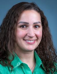 Headshot of smiling woman with curly brown hair wearing a green collared shirt, blue background
