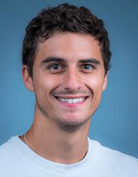 Headshot photo of smiling young man with dark curly hair against blue background