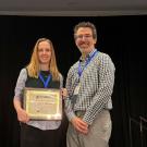 Two smiling people on stage holding a framed award plaque