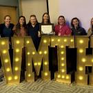 Ten people smiling behind large illuminated 'VMTH' marquee letters, one holding a plaque