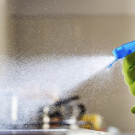 A gloved hand sprays cleaning solution, creating a mist in a bright kitchen.