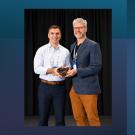 Two men in smart casual attire smiling on stage; younger man holds an award